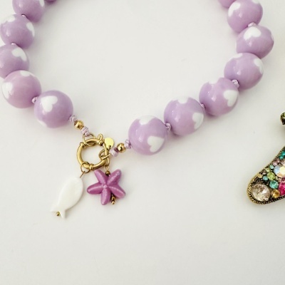 Pulseira com contas lilás e corações brancos, fecho dourado e pendentes decorativos