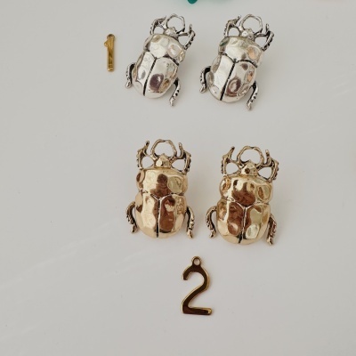 Brincos em forma de escaravelho dourados e prateados com números 1 e 2 dourados em fundo branco.
