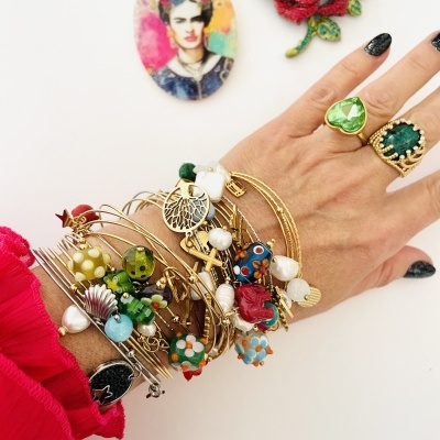 Mão com pulseiras douradas coloridas e anéis verdes, fundo branco com imagens de Frida Kahlo e rosa vermelha