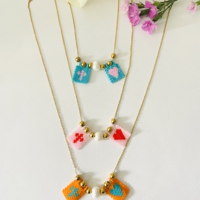 Três colares dourados com pendentes de contas coloridas e pérolas, sobre superfície branca com flores roxas ao lado.