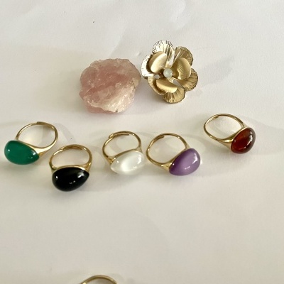 Seis anéis dourados com pedras coloridas sobre fundo branco, com pedra rosa e flor dourada ao fundo