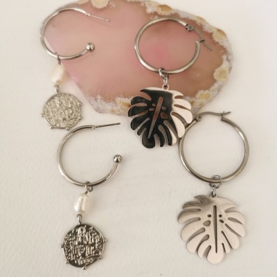 Quatro brincos de argola prateada com pendentes de folha, moeda e pérola sobre base rosa e flor branca