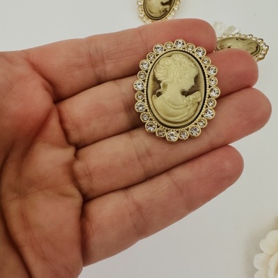 Broches dourados com perfil feminino em relevo segurados por mão sobre fundo branco.