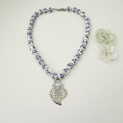 Colar com contas brancas e padrão azul com pendente prateado, ao lado duas flores decorativas
