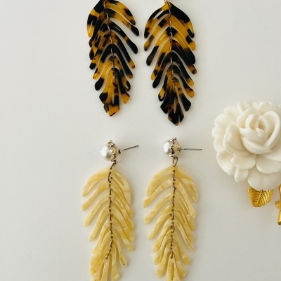 Dois pares de brincos pendentes em forma de folhas, um castanho e preto e outro bege perolado, sobre fundo branco com flor decorativa.
