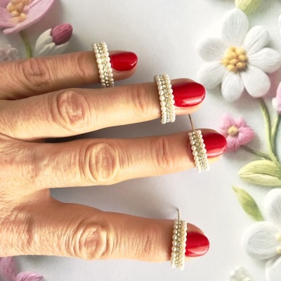 Mão com quatro anéis de pérolas brancas em dedos com unhas vermelhas sobre fundo floral branco.