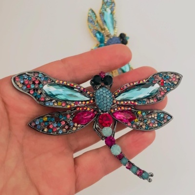 Broche de libélula com pedras coloridas, segurado por uma mão.