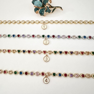 Quatro pulseiras numeradas de metal dourado com pedras coloridas e broche de flor verde sobre fundo branco.