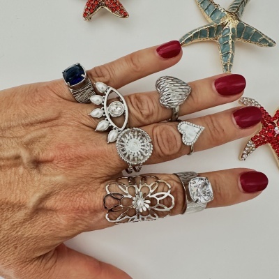 Mão com vários anéis prateados e unhas vermelhas, fundo branco com estrelas do mar decorativas.