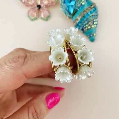 Anel dourado com flores brancas, broches borboleta rosa e concha azul ao fundo