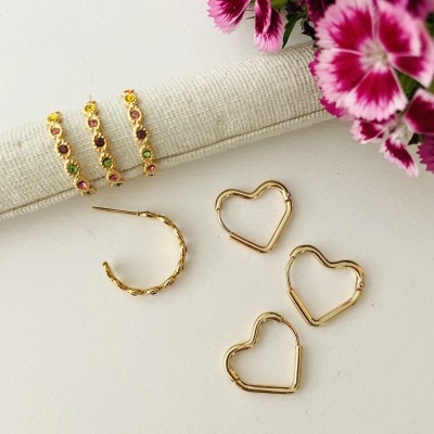 Brincos de metal dourado com pedras coloridas e em forma de coração junto a tecido creme e flores roxas.