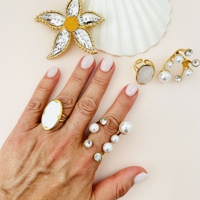 Anel e brincos dourados com pérolas e pedras transparentes junto a mão com unhas brancas e concha branca