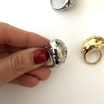 Três anéis largos com pedras coloridas, um sendo segurado por mão com unha vermelha.