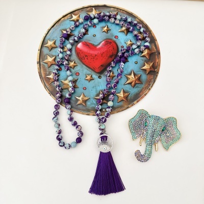 Colar de contas roxas e azul claro com tassel roxo e broche de elefante com pedras azuis.