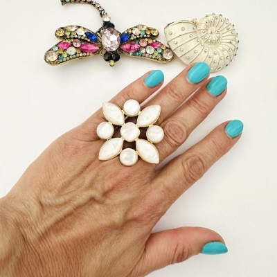 Anel grande dourado com pedras brancas em mão com unhas azuis, junto a dois broches coloridos sobre fundo branco.