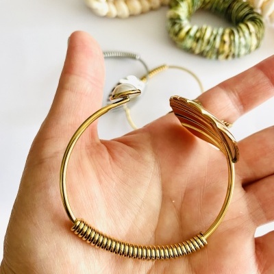 Pulseira dourada com molas nas extremidades, segurada por mão, com pulseiras coloridas ao fundo