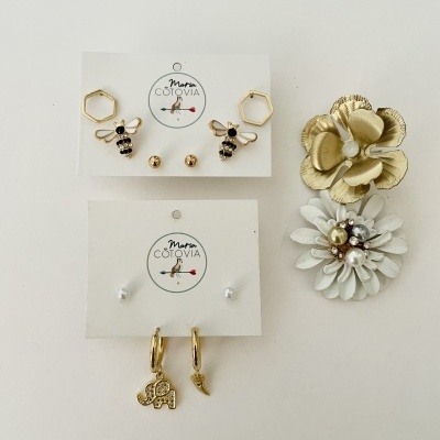 Conjunto variado de brincos dourados e brancos com abelhas, hexágonos, pérolas, elefante e pena sobre fundo branco.