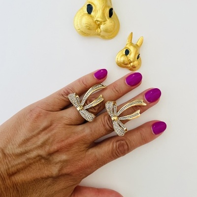 Anéis dourados com laços em mão com unhas roxas e duas figuras douradas de coelhos.
