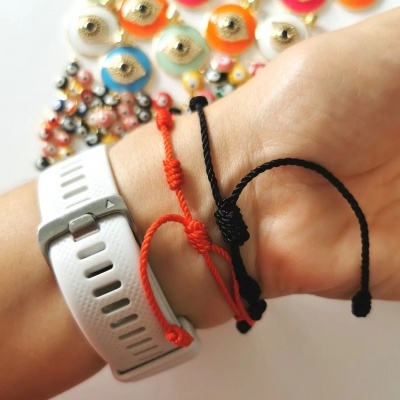 Pulseiras de cordão vermelha e preta no pulso ao lado de bracelete branca, com pulseiras coloridas em fundo