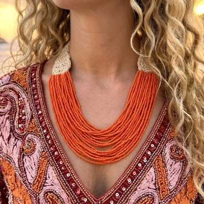 Colar de contas laranja multifio com fecho em croché bege no pescoço de uma mulher com vestido bordado colorido