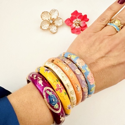 Pulseiras e anel esmaltados com flores coloridas no braço de uma pessoa com duas flores decorativas ao fundo
