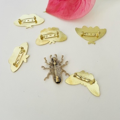 Pins dourados em formato de folhas e aranha com fecho de alfinete, sobre fundo branco ao lado de flor rosa