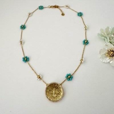 Colar dourado com medalhão central e flores azuis e brancas sobre fundo branco
