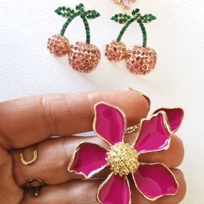 Brincos decorativos com pedras e esmalte, formato de cereja e flor, sobre fundo branco.