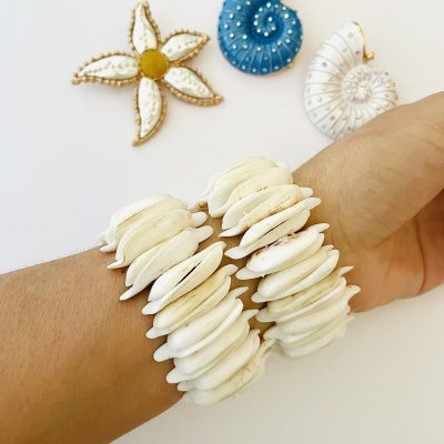 Pulseira de conchas naturais brancas com broches decorativos em formato de flor e caracóis ao fundo