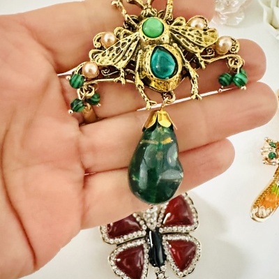 Broche dourado com pedras verdes e pérolas, pendente em gota verde, sobre mão, fundo branco.