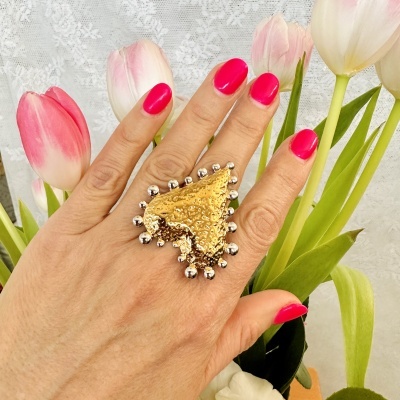 Anel dourado texturado com bolas prateadas, mão com unhas pintadas de pink, tulipas cor-de-rosa e brancas ao fundo.