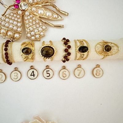 Anéis dourados com pedras negras e vermelhas numa faixa branca ao lado de um broche dourado decorado com pérolas e pedras brilhantes.