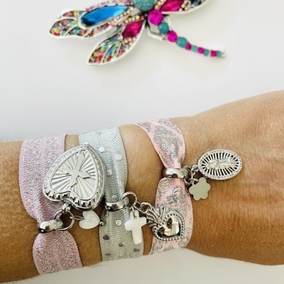 Pulseira elástica rosa e cinza com medalhões metálicos e crucifixo no pulso, libélula colorida ao fundo