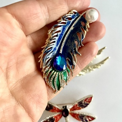 Broche de pena colorida com pedra azul e pérola, mão a segurá-lo, broche de libélula ao fundo