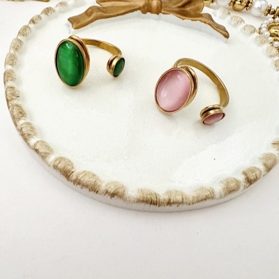 Dois anéis dourados com pedras verde e rosa sobre prato branco com borda dourada e colar perolado ao fundo