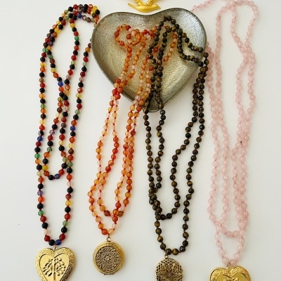 Quatro colares coloridos com pendentes dourados dispostos junto a um prato em forma de coração cinza e adorno dourado.