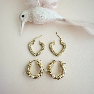 Dois pares de brincos dourados, um com formato de coração e pedras, outro com textura e pérolas, e um pássaro brilhante branco como adorno