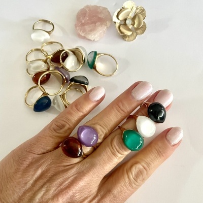 Anéis dourados com pedras coloridas usados numa mão com unhas pintadas e mais anéis e pedras numa superfície branca