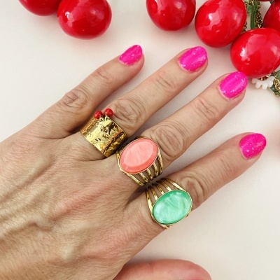 Mão com três anéis dourados com pedras rosas e verdes, unhas cor de rosa brilhante e cerejas artificiais vermelhas ao fundo