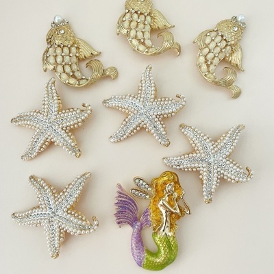Bijuterias com tema marinho incluíndo peixes dourados, estrelas-do-mar peroladas e sereia colorida sobre fundo bege.