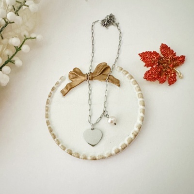 Colar com coração prateado e pérola em prato branco decorado com laço castanho e folha vermelha decorativa