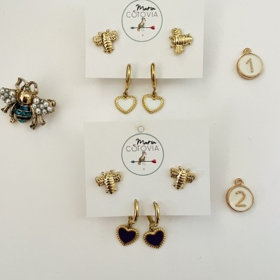 Brincos dourados em cartão branco da Maria Cotovia, com desenhos de abelhas e corações, e medalhões numerados 1 e 2 mais um broche de abelha com pérolas.