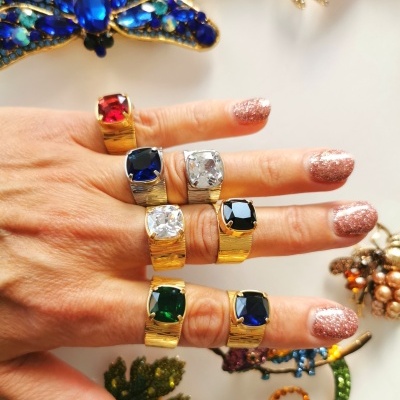 Mão com anéis de pedras coloridas e unhas cintilantes sobre fundo branco com joias de libélula e folhas.