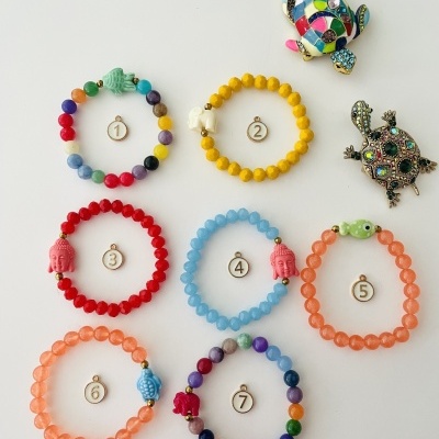 Sete pulseiras coloridas numeradas com contas redondas e duas tartarugas decorativas ao lado.
