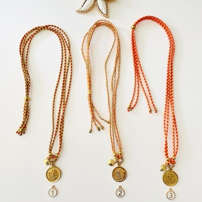Três colares trançados laranja com pendentes dourados numerados 1 a 3 e flor decorativa branca com centro amarelo sobre fundo branco