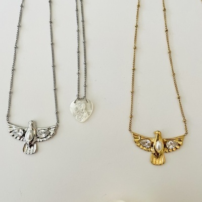 Três colares com pendentes de pássaro dourado e prateado, e um de coração prateado, sobre fundo branco