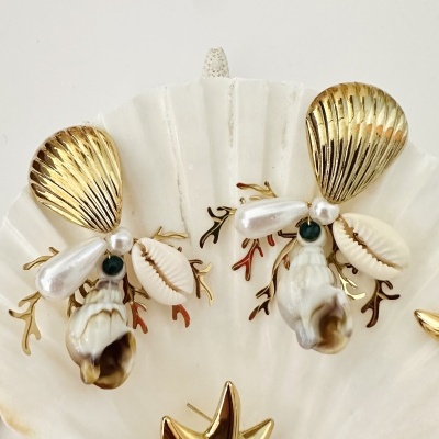 Brincos decorativos com conchas, peças douradas e pérolas sobre concha branca maior
