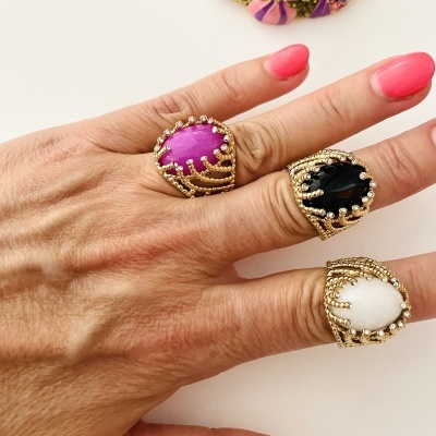 Mão feminina com três anéis grandes coloridos e uma máscara decorativa ao fundo