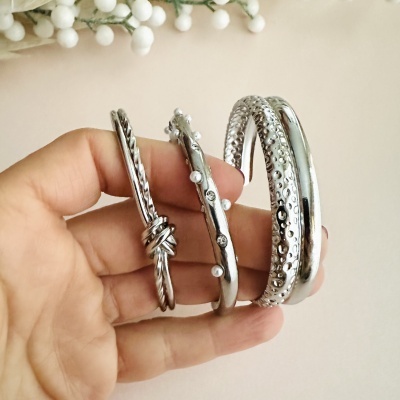 Conjunto de pulseiras prateadas com texturas e pérolas, seguradas por mão sobre fundo bege.