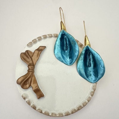 Brincos azuis florais com base dourada sobre prato branco decorado com laço castanho.